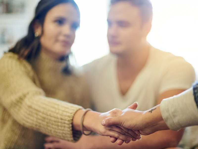 Two people clasp hands with their counselor in a warm, sunlit room during a supportive counseling session.