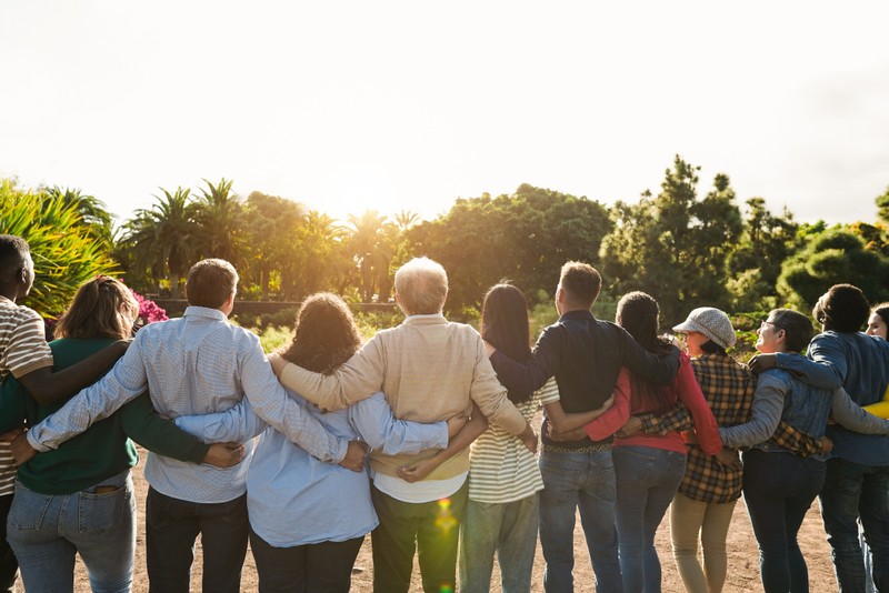 A diverse group of people embrace outdoors at golden hour, arms around one another in a moment of community support.